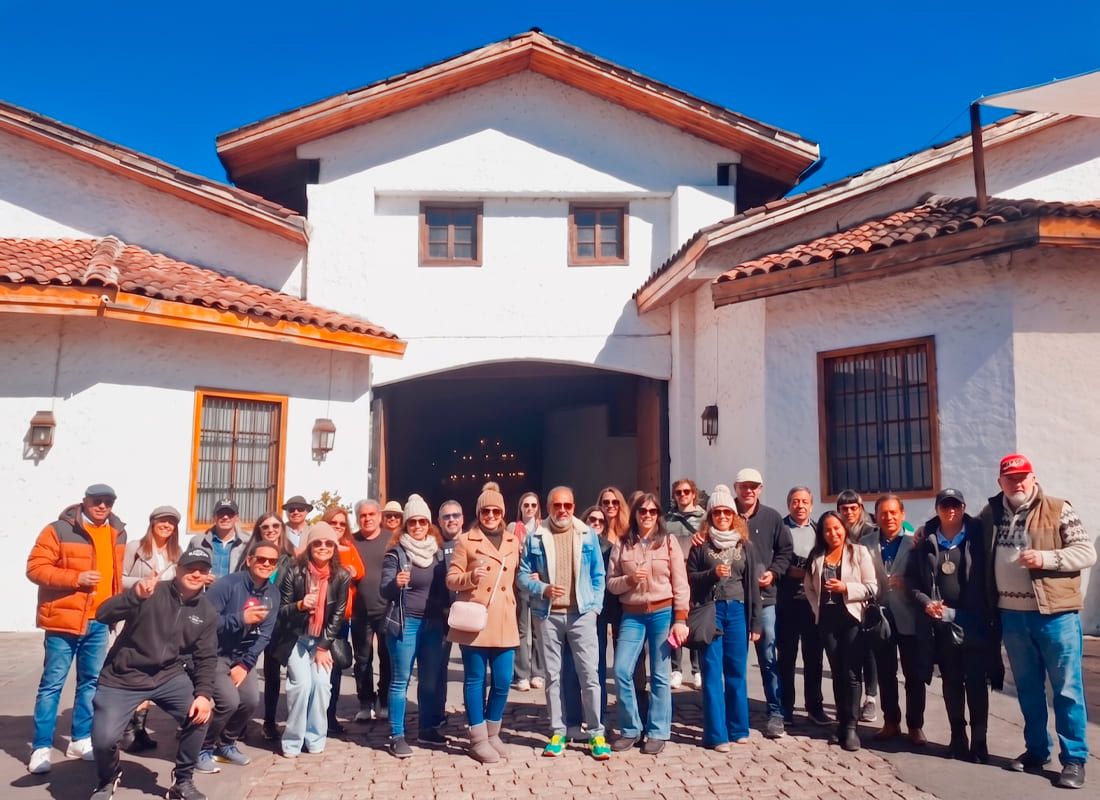 Fotografía diurna de un grupo grande de turistas sonriendo y posando sobre un suelo de adoquines, varios de ellos sosteniendo copas de vino de degustación. Se encuentran frente a la fachada principal de una tradicional casona blanca de estilo colonial con techo de tejas. Detrás del grupo destaca un gran portón abierto que revela un interior de bodega con iluminación cálida, todo bajo un cielo azul despejado.