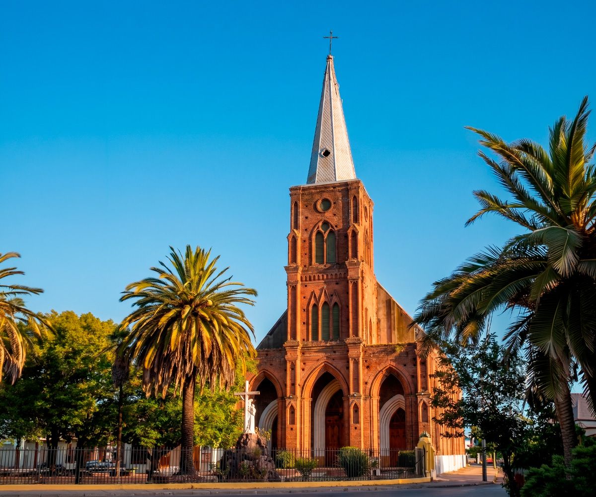 Fotografía de la fachada de una histórica iglesia de ladrillo rojo con una gran torre campanario central, rodeada de palmeras y vegetación en Curicó, Chile, bajo un cielo azul despejado.