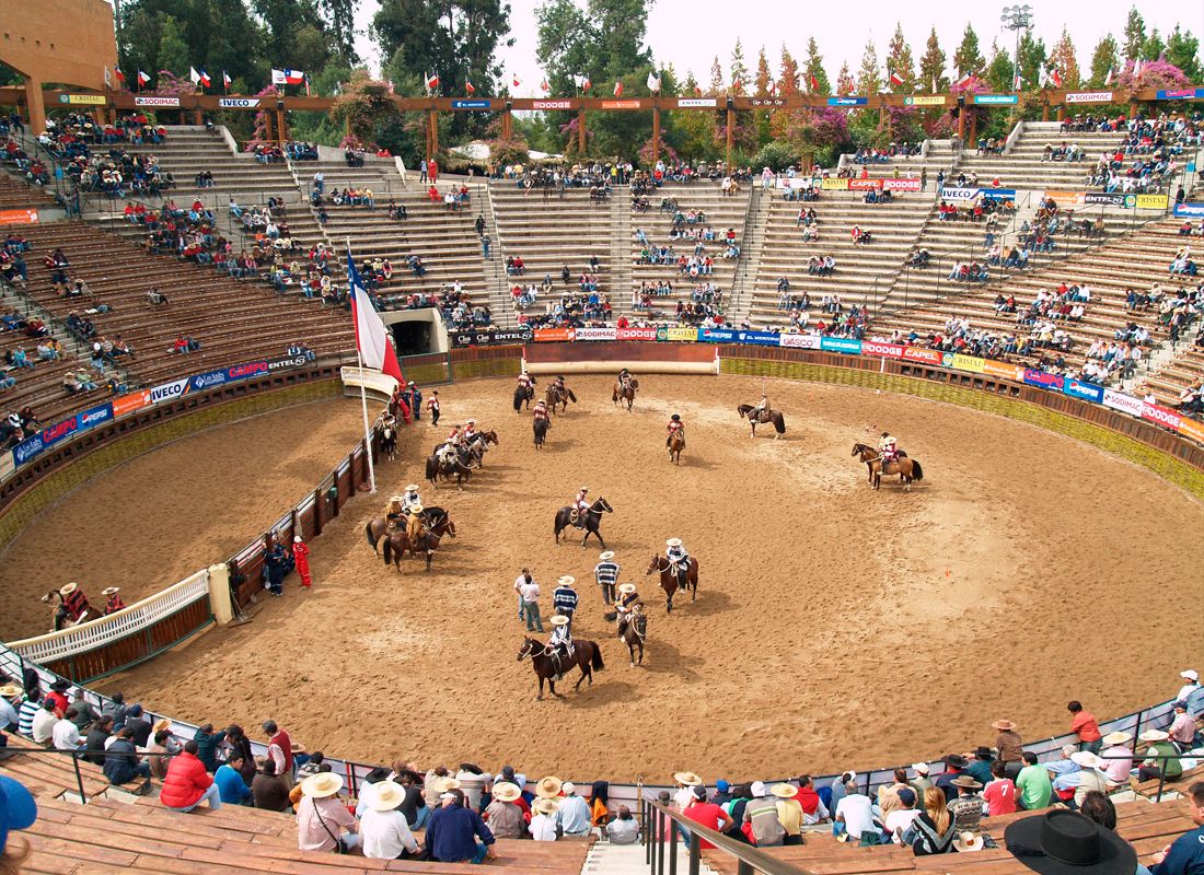 Fotografía panorámica del interior de la Medialuna Monumental de Rancagua durante un evento. En la arena central, varios jinetes con vestimenta tradicional de huaso chileno montan a caballo, destacando uno que sostiene una gran bandera de Chile. Las graderías del recinto están llenas de espectadores observando la presentación, rodeados de carteles publicitarios y árboles en la parte superior.