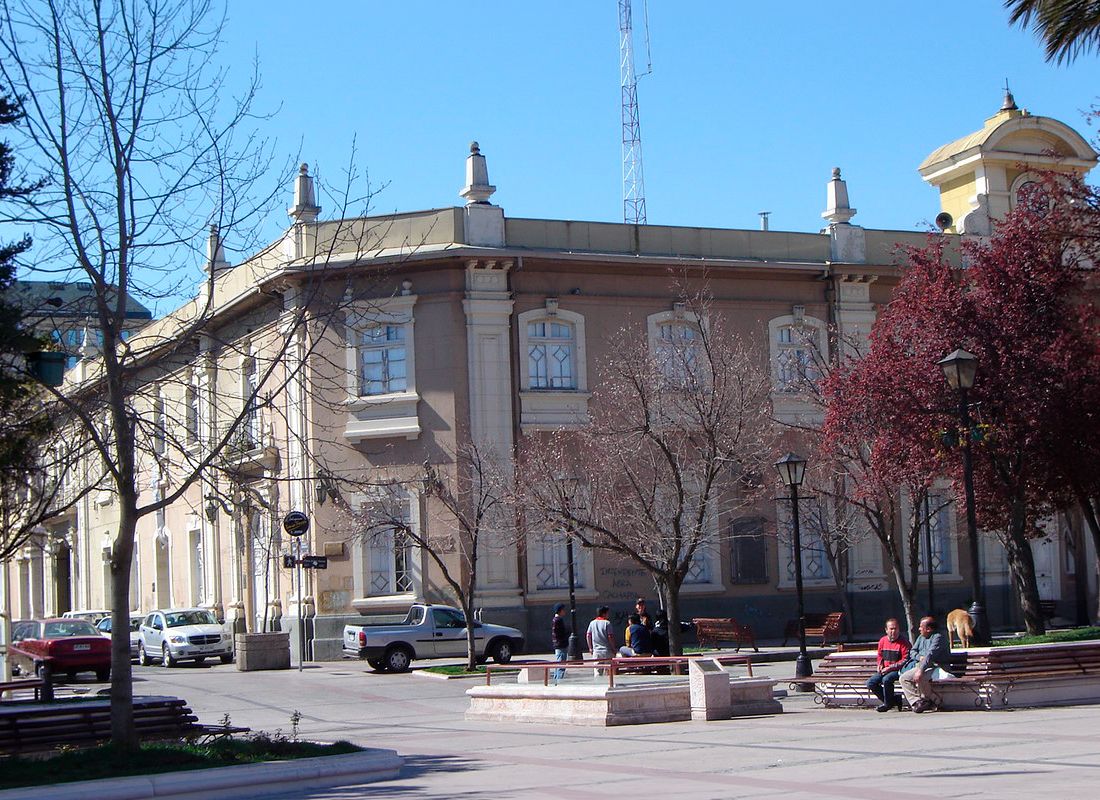 Fotografía diurna de una plaza en Rancagua. En primer plano se observan personas conversando y descansando en bancas de madera, rodeadas de árboles de hoja caduca y senderos pavimentados. De fondo, destaca un edificio institucional de dos pisos y estilo clásico, con fachada color beige, grandes ventanas blancas en arco y detalles arquitectónicos en el techo.