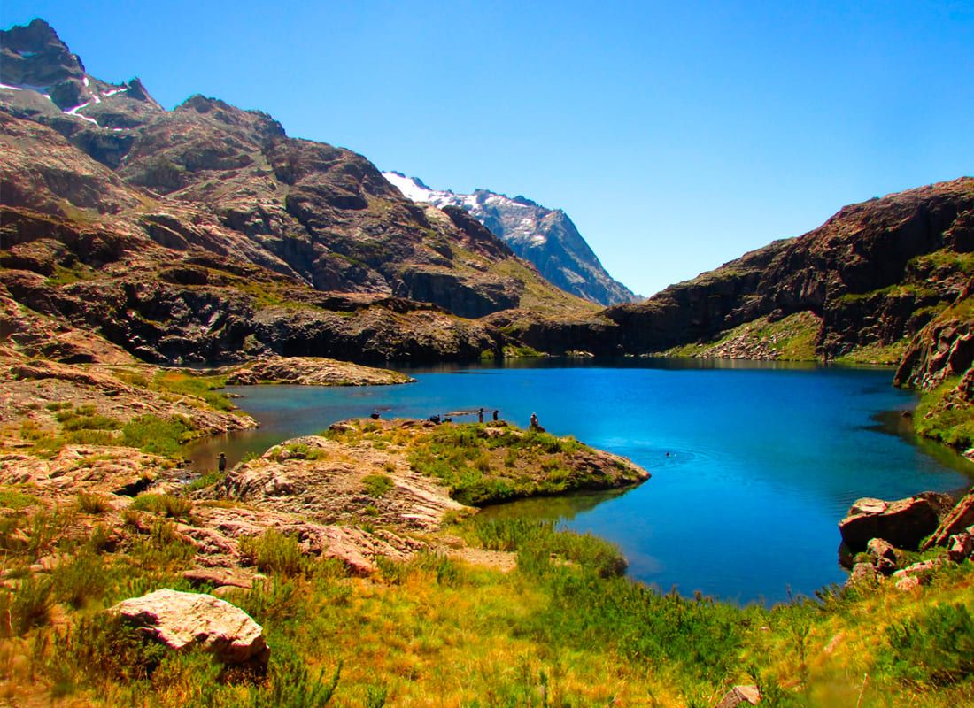 Fotografía panorámica de un impresionante paisaje natural en la precordillera. Destaca una hermosa laguna de aguas cristalinas y de color azul intenso, rodeada por montañas rocosas y laderas con vegetación verde y amarillenta, bajo un cielo despejado. Representa los atractivos vírgenes de la zona de Linares, como el Cajón del Río Achibueno.