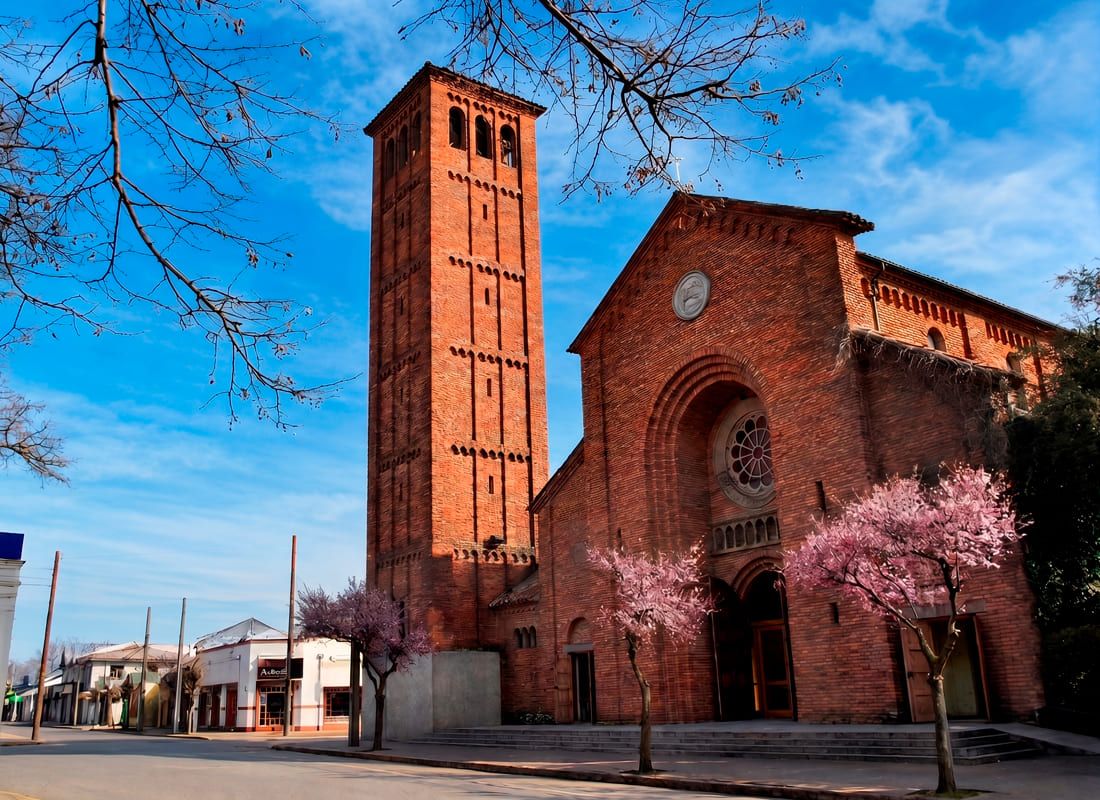 Fotografía diurna de la Catedral de San Ambrosio en el centro de Linares. Destaca su imponente construcción de ladrillos rojos con una alta torre campanario, contrastando bajo un cielo azul intenso. En la calle peatonal frente a la fachada se aprecian árboles con flores rosadas en floración.
