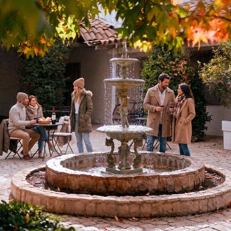 Fotografía de un patio colonial pavimentado con adoquines, destacando una gran fuente de agua clásica de piedra con varios niveles en el centro. Alrededor, dos parejas con ropa de abrigo de temporada otoño-invierno conversan sonrientes mientras disfrutan de copas de vino tinto. La escena está enmarcada en la parte superior por hojas otoñales, y de fondo se aprecia la arquitectura tradicional de una casona con techo de tejas.