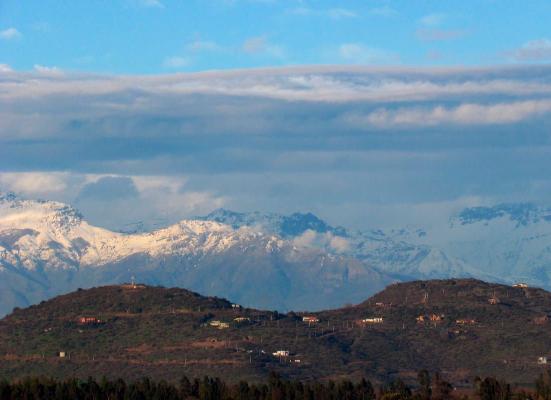 Fotografía de paisaje diurno. En el fondo destaca la imponente Cordillera de los Andes cubierta de nieve bajo un cielo azul con nubes horizontales. En el plano medio se observan cerros más bajos con vegetación oscura y algunas casas dispersas, mientras que en la base de la imagen hay una densa línea de árboles.