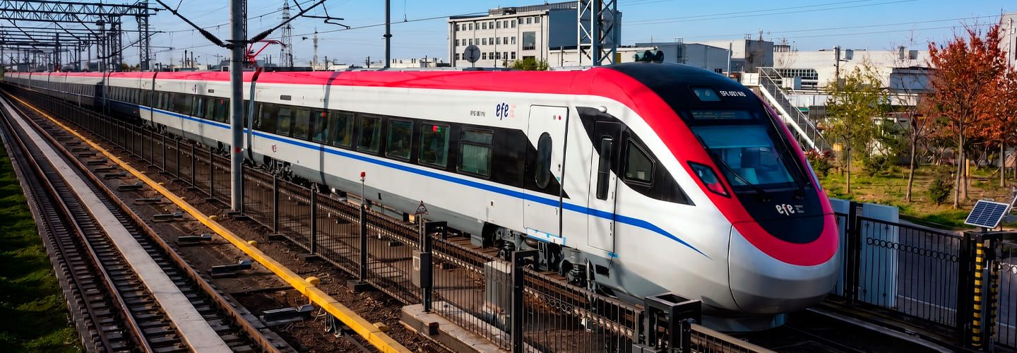 Fotografía en formato panorámico de un moderno tren bimodal (BMU) de EFE avanzando por las vías férreas. Destaca el diseño aerodinámico y el brillante color rojo y blanco de la cabina y los vagones. El convoy ferroviario se desplaza a gran velocidad durante su trayecto de conexión hacia el sur.