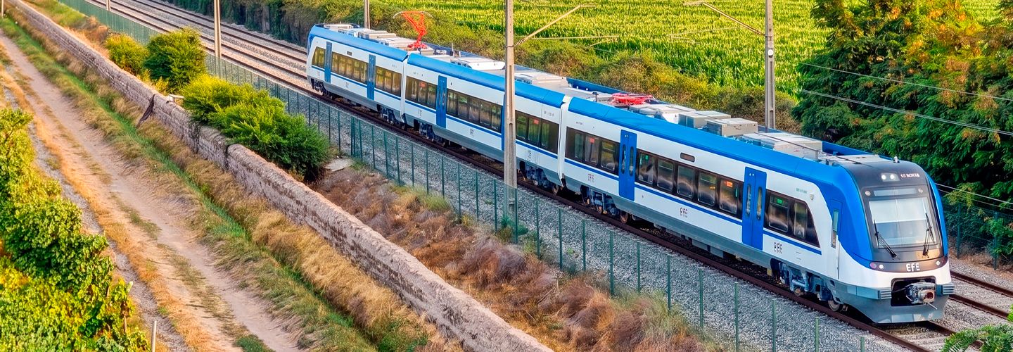 Vista panorámica media de un tren de pasajeros moderno de la empresa EFE (Empresa de los Ferrocarriles del Estado) para el servicio Santiago-Rancagua, de color azul claro y blanco, circulando por vías férreas sobre un terraplén elevado con árboles detrás.