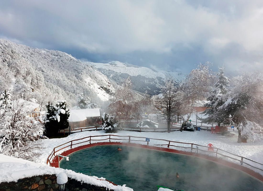 Fotografía de una piscina termal al aire libre en Nevados de Chillán, rodeada de un paisaje de montañas cubiertas de nieve bajo un cielo parcialmente nublado. Imagen visible en la imagen