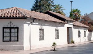 Fotografía diurna de la fachada exterior del Museo Regional de Rancagua. Destaca una casona tradicional de estilo colonial de un solo piso, con paredes de adobe pintadas de blanco, techo de tejas de arcilla y ventanas protegidas por herrería artística. El edificio se encuentra junto a un paseo peatonal pavimentado bajo un cielo azul despejado.