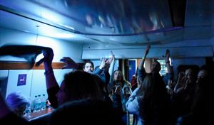 Fotografía del interior de un vagón de tren turístico ambientado como discoteca. En la oscuridad, un grupo de personas baila animadamente y levanta las manos, iluminados por luces azules de fiesta. La escena refleja el ambiente festivo del 'discotren' durante el viaje de regreso.