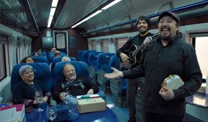 Fotografía del interior de un vagón de pasajeros en un tren turístico. En el pasillo central, músicos interactúan alegremente con los pasajeros sentados en las butacas azules, ofreciendo un espectáculo de música en vivo a bordo durante el trayecto.